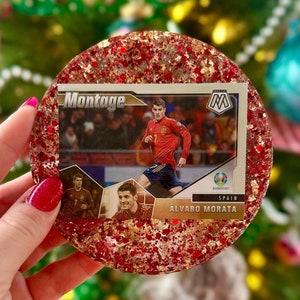 May include: A red and gold glitter resin coaster with a soccer trading card featuring a player in a red and yellow jersey. The card reads "Montage" and "Álvaro Morata".