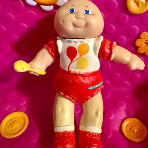 May include: A vintage Cabbage Patch Kids doll with a bald head, blue eyes, and a smiling expression. The doll wears a white shirt with red and yellow balloons, red shorts, and red shoes. It holds a yellow toy and is surrounded by orange plastic pieces.