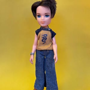 May include: A Bratz doll with brown hair and blue eyes, wearing a mustard yellow and navy blue top with a dragon design, blue jeans, and brown shoes. The doll is posed against a yellow background.