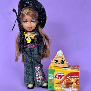 May include: A Bratz doll wearing a black hat, denim jacket, and jeans with floral embroidery. Next to the doll is a Shopkins waffle toy with a box of Kellogg's Eggo Homestyle waffles.