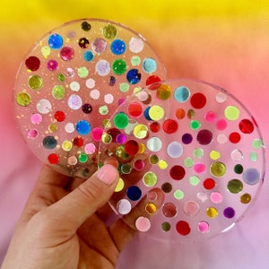 May include: Two round clear resin coasters with colorful confetti embedded inside. The coasters are translucent and show the confetti through the resin.