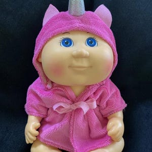 May include: A doll with a round face, blue eyes, and rosy cheeks, wearing a bright pink hooded robe. The hood features two small ears and a central silver, glittery unicorn horn. The robe is made of a soft, plush fabric and ties at the front.