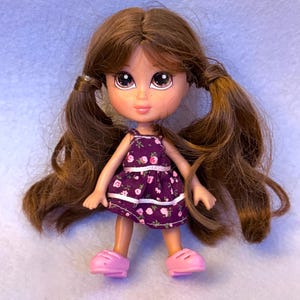 May include: A Bratz doll with brown hair styled in pigtails. The doll has large eyes and is wearing a purple dress with a floral pattern and pink shoes. The doll is a collectible toy.