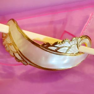 May include: A decorative, curved object with a pearlescent finish, accented with gold trim and leaf-like details. The object has a central opening and is set against a pink background. The design is ornate and elegant.