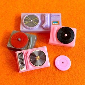 May include: A collection of miniature toy turntables in various colors, including pink, gray, and purple, with black and red vinyl records. The turntables are arranged on an orange surface.