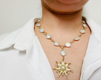 Peut inclure: Un collier doré avec un pendentif en forme de soleil, un diamant central et des cristaux environnants. Le collier est composé d'une chaîne de perles blanches irrégulières et de maillons dorés. Le collier est porté sur une chemise blanche.