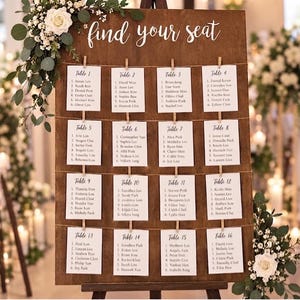 May include: A dark wood wedding seating chart sign featuring the white text find your seat, decorated with white roses and eucalyptus. Sixteen white paper cards are clipped to horizontal twine strings, each listing table numbers and guest names.