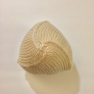 May include: A beige knitted hat with a ribbed texture. The hat is made of a soft yarn and has a rounded shape. The hat is designed to fit snugly on the head and is suitable for cold weather.