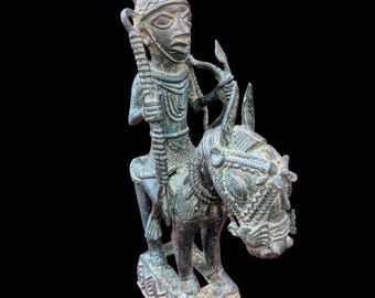 Pode incluir: Estátua de bronze que retrata uma figura montada num animal, possivelmente um cavalo ou um burro. A figura usa uma coroa e segura um bastão. A estátua tem detalhes intrincados e é montada numa base com padrões geométricos. A estátua é de cor bronze escuro.