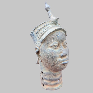 Könnte beinhalten: Eine Bronzeskulptur eines menschlichen Kopfes mit detaillierter Krone und Gesichtszügen. Der Kopf hat einen ruhigen Ausdruck und ist mit komplizierten Mustern und Texturen verziert. Die Skulptur ist ein historisches Artefakt.