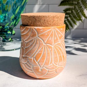 May include: A peach-colored ceramic jar with a carved, repeating fan-like pattern across its exterior surface. The jar is topped with a cylindrical, light brown cork lid that fits securely into the opening of the vessel.