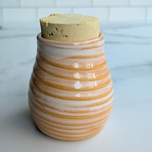 May include: A small, white ceramic jar with a cork lid. The jar has a striped pattern in shades of orange and white.