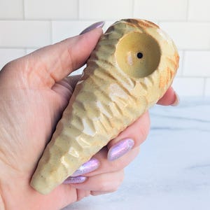 May include: A ceramic pipe with a textured, cone-like shape. The pipe is primarily beige with brown and orange accents. The bowl is circular with a small hole in the center.