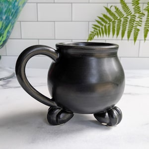 May include: A black ceramic mug with a rounded body and a large handle. The mug sits on three small, rounded feet. The mug has a glossy finish and is likely handmade. The mug is reminiscent of a witch's cauldron.