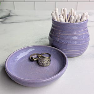 May include: A lavender ceramic jar with horizontal ridges filled with white cotton swabs, accompanied by a matching lavender ceramic dish containing three metal rings, including one with a decorative filigree pattern and a dark bead.