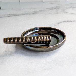 May include: A small, round, ceramic ashtray with a built-in rest. The ashtray is dark with a metallic sheen, and the rest is a ridged, cylindrical shape. The ashtray is designed for holding cigarettes or cigars.