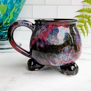 May include: A handmade ceramic mug with a unique, colorful glaze. The mug features a dark red handle and a body with a blend of red, blue, purple, and black hues. It rests on three black, rounded feet, adding a distinctive touch.