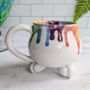 May include: A white ceramic mug with a large handle and three small feet. The rim and sides of the mug are decorated with colorful drips of paint in shades of orange, red, purple, blue, and green. The mug is sitting on a white surface.