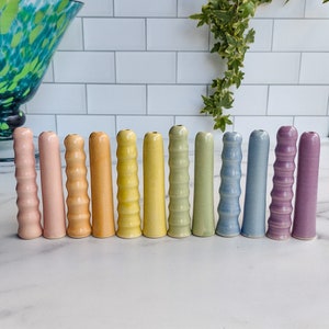 May include: A set of 12 ceramic finger pots in a rainbow of pastel colors. The pots are arranged in a row from left to right, with the colors gradually transitioning from light pink to purple.