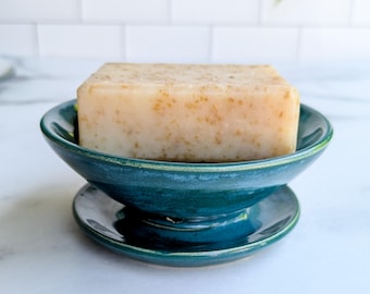 May include: A rectangular bar of beige soap with visible flecks of botanical material resting inside a deep teal ceramic soap dish. The soap dish features a wide, rounded bowl shape with a matching circular base, showcasing a glossy finish.