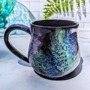 May include: A ceramic mug with a glossy finish featuring a swirling nebula-like pattern of deep black, vibrant purple, electric blue, and bright green. The mug has a rounded body and a curved handle, with a smooth, dark interior and a wide rim.