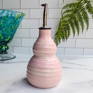 May include: A pink ceramic oil bottle with a stainless steel spout. The bottle has a bulbous shape with horizontal textured lines. The spout is long and slender. The background includes a green glass bowl and a fern.