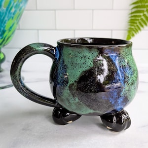 May include: A handmade ceramic mug with a unique design. The mug features a glossy finish with a blend of green, blue, and black colors. It has a rounded body, a large handle, and is supported by three small, claw-shaped feet.