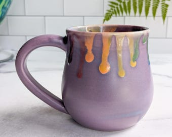 May include: A handcrafted ceramic mug in a soft purple hue, featuring a unique design with colorful drips of glaze around the rim. The mug has a large handle and a smooth, glossy finish, perfect for enjoying your favorite beverage.