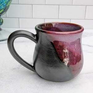 May include: A ceramic mug featuring a rounded body with a smooth matte black finish on one side and a glossy, deep magenta-purple glaze on the other. The mug includes a curved black handle and a wide, open rim with a dark interior.