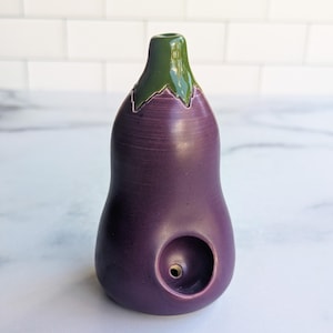 May include: A ceramic eggplant-shaped pipe. The body is a deep purple, with a green stem and a small hole on the side. The pipe is designed to resemble a realistic eggplant.