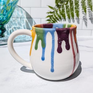 May include: A white ceramic mug with a rounded body and a large handle, featuring a colorful drip pattern around the rim. The glaze drips down the sides in vibrant shades of yellow, green, blue, purple, and red, creating a smooth, glossy finish.