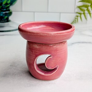 May include: A pink ceramic wax warmer with a crescent moon cutout. The warmer has a round dish on top and a cylindrical base. The color is a muted, dusty rose, and the surface has a textured appearance. The design is simple and elegant.