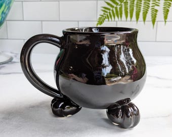 May include: A glossy black ceramic mug with a rounded body and a large, curved handle. The mug is supported by three small, pumpkin-shaped feet. The mug's surface reflects the surrounding environment.