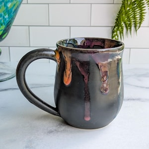 May include: A handmade ceramic mug with a black exterior and a dark interior. The mug features a rounded shape with a large handle. The glaze has a glossy finish with drips of orange, purple, and red. The mug is approximately 4 inches tall.