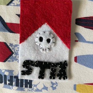 Può includere: Un cerotto di feltro rosso e bianco con un design di teschio e ossa incrociate e la parola "STIX" ricamata in filo nero.