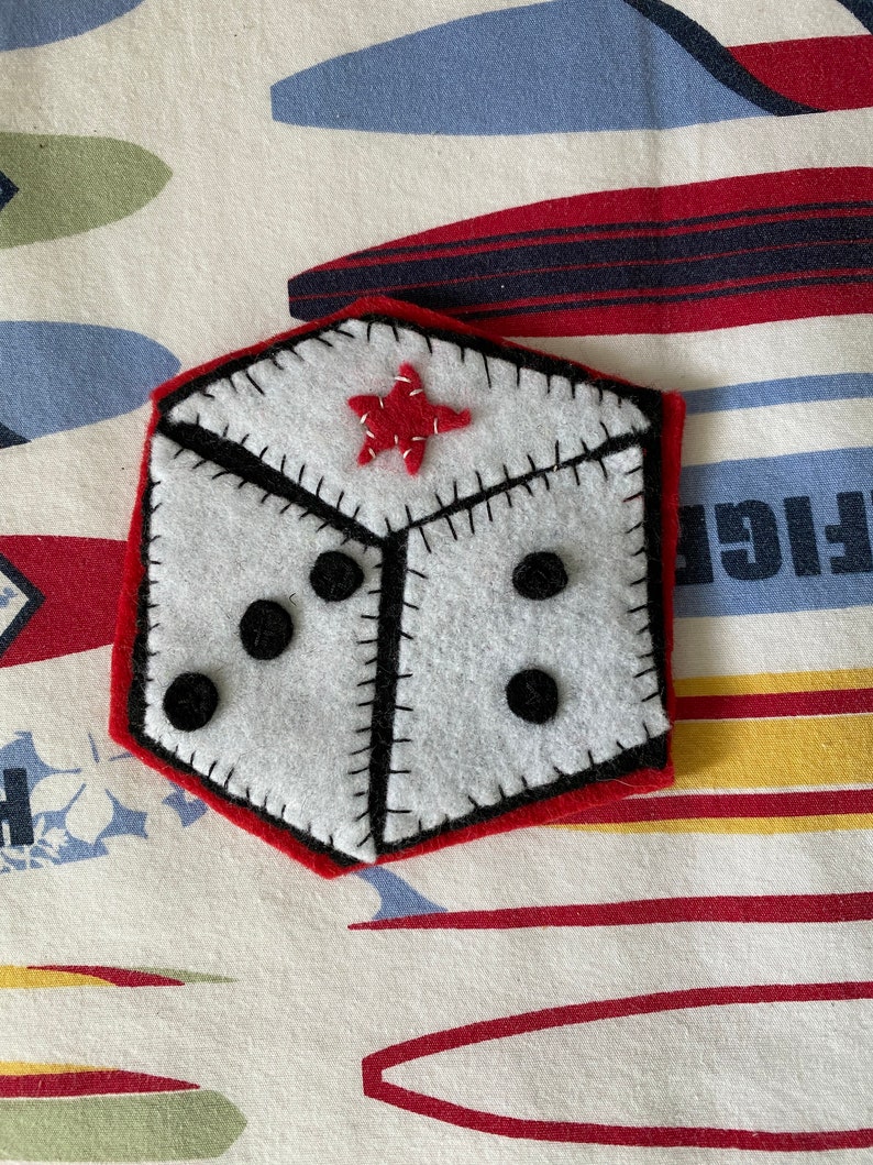 May include: A white felt dice with black dots and a red felt star on one side. The dice is sewn with black thread and has a red felt border.