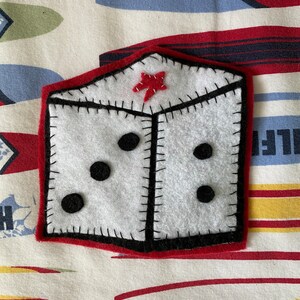 May include: A white felt dice with black dots and a red felt star on top. The dice is sewn with black thread and has a red felt border.
