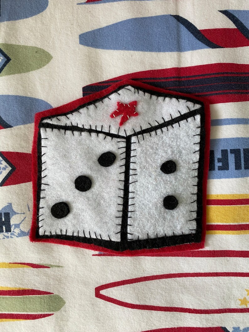 May include: A white felt dice with black dots and a red felt star on top. The dice is sewn with black thread and has a red felt border.