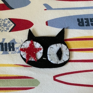 May include: A black felt cat with a red star on one eye and a white felt eye on the other. The cat is on a white background with a pattern of colourful surfboards.