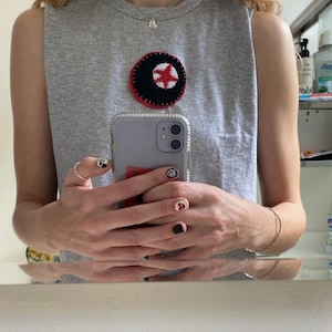 Pode incluir: Uma camiseta cinza sem mangas com um remendo bordado em vermelho, preto e branco na frente. A pessoa está usando um colar de prata e um anel de prata no dedo. A pessoa está segurando uma capa de telefone preta com um telefone branco dentro.