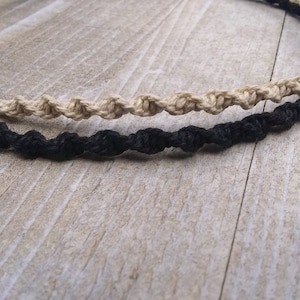 May include: Two crocheted necklaces, one black and one beige, are displayed on a wooden surface. The necklaces are made with a simple knot pattern.