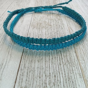 May include: Two handmade macrame bracelets in shades of teal and turquoise. The bracelets are made of woven cord and are displayed on a light wood surface. The bracelets are adjustable and have a simple, minimalist design.