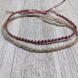 May include: Two friendship bracelets, one brown and pink, the other natural beige, are tied together and laid out on a white wooden surface.