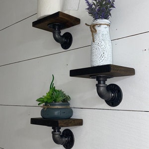 May include: Three rustic wooden shelves with black metal pipe brackets mounted on a white wall. The top shelf holds a roll of toilet paper, the middle shelf holds a white vase with purple flowers, and the bottom shelf holds a small potted succulent.