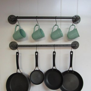 May include: A kitchen storage rack with two horizontal black metal bars. Four teal mugs hang from the top bar, and four black frying pans hang from the bottom bar. The rack is mounted on a white wall.
