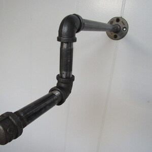 May include: Black metal pipe shelf with a 90-degree bend and a flange on the wall mount.