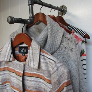 May include: A black metal pipe clothing rack with three wooden hangers holding a striped sweater, a grey knitted sweater, and a striped shirt with a "Luii" label.