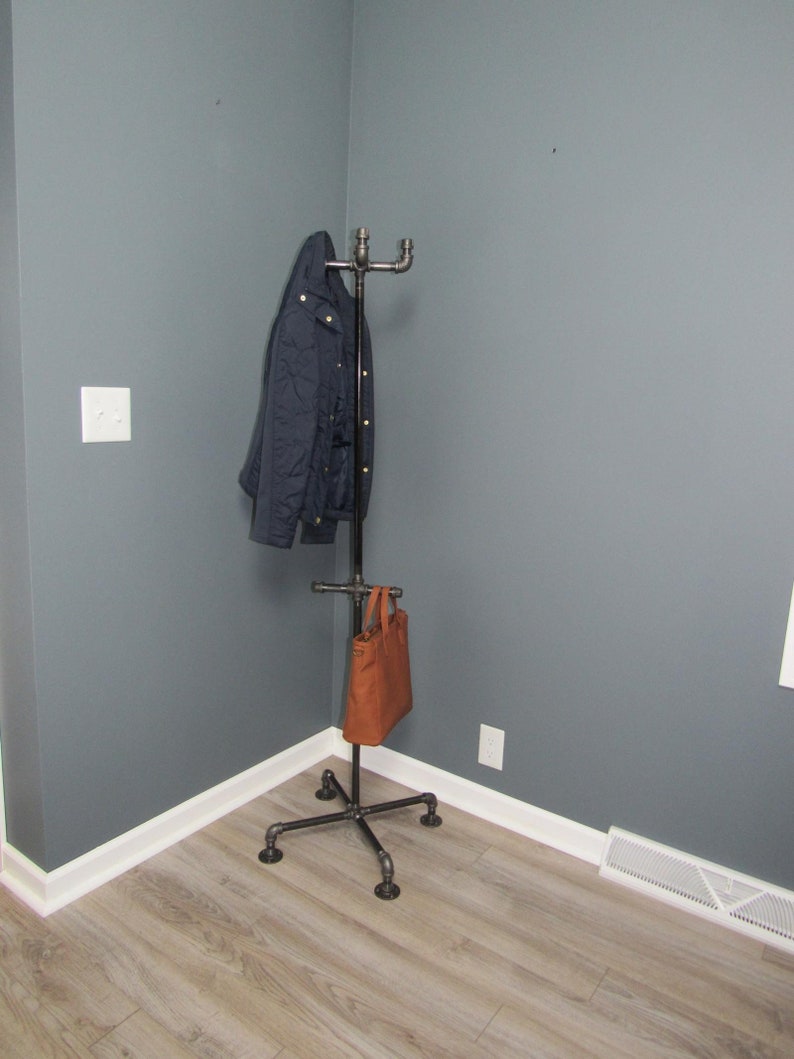 May include: A black metal coat rack with a brown leather bag hanging on it. The rack is in the corner of a room with gray walls and white trim.
