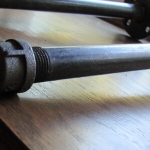 May include: Close-up of a black metal pipe fitting with a threaded end. The fitting is attached to a black metal pipe. The pipe is lying on a wooden surface.