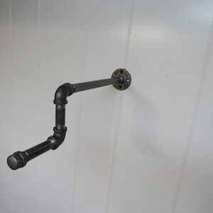 May include: Black metal pipe shelf with a flange mounted to a white wall. The shelf is bent into an L-shape.